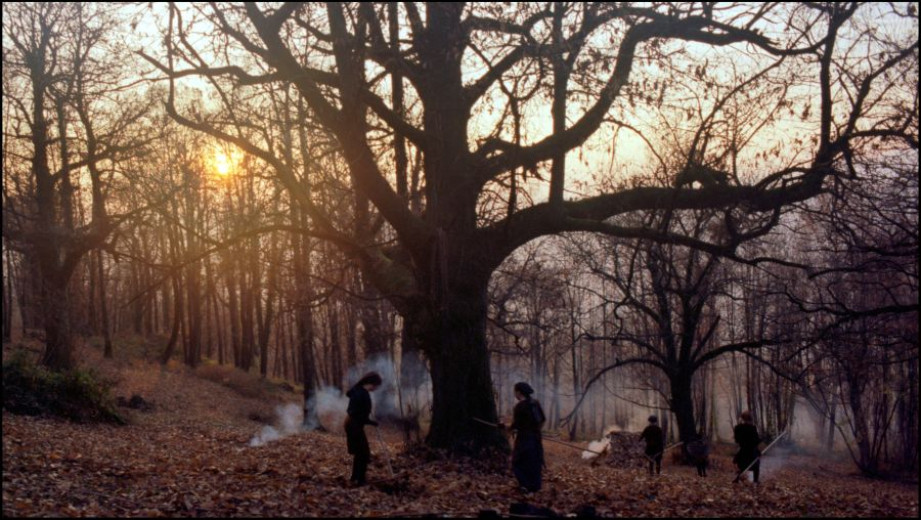 A photo of an autumn forest with the sun shining in the background. Four people dressed in dark clothing are holding rakes, and there is smoke visible nearby.