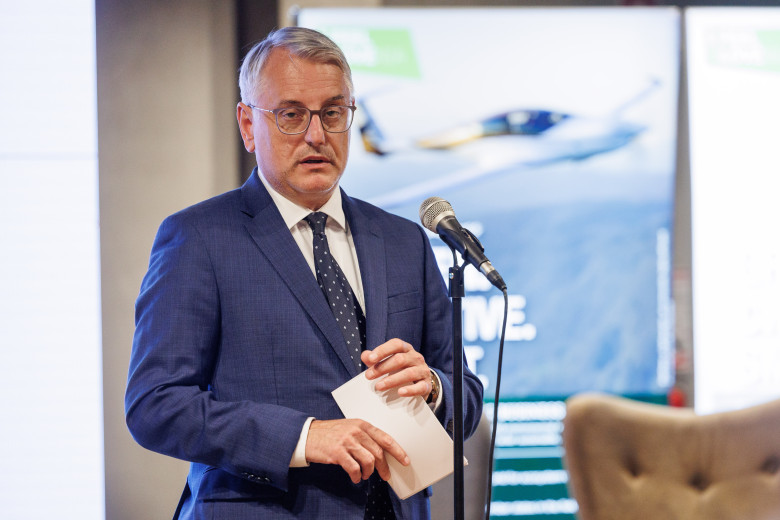 Matjaž Han standing behind a microphone, pictured up to his waist, with a photo billboard in the background.