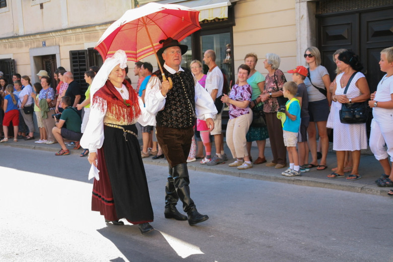 A couple in Gorenjska national costume walks in a procession under an umbrella, with event visitors in the background.