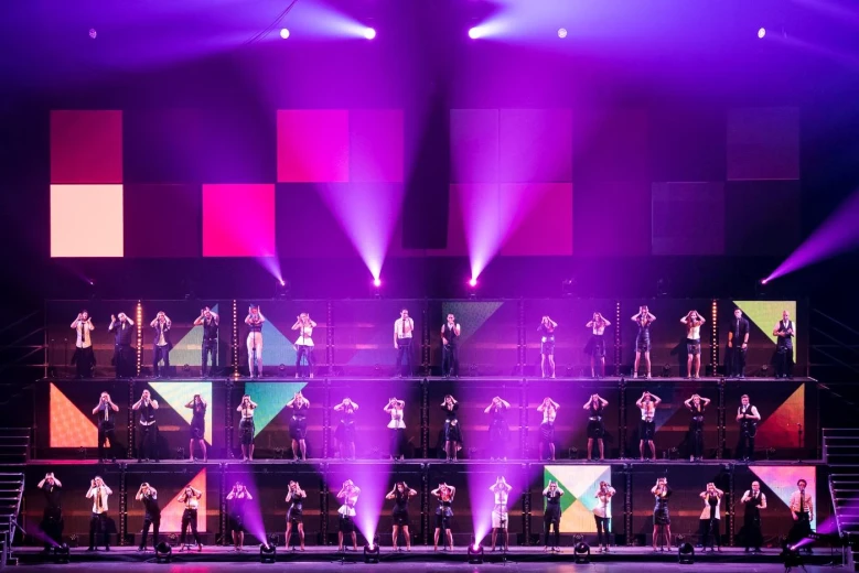 Perpetuum Jazzile singers are performing on stage in rows that are separated into boxes on three levels. They are illuminated by purple, pink and orange lights.