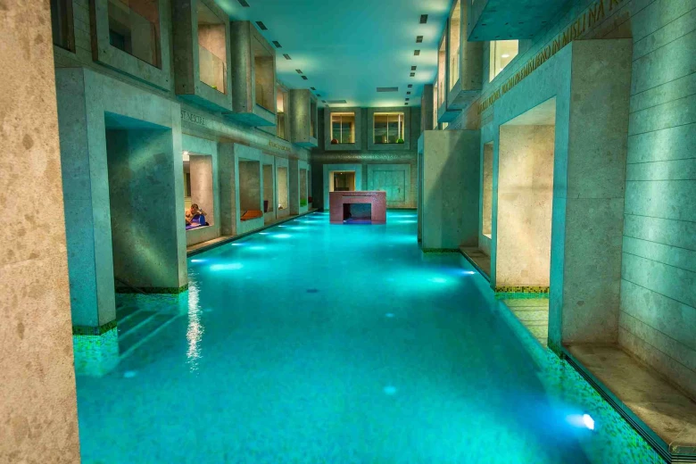 The indoor pool of the Roman Baths with calm turquoise water in a large space surrounded by stone columns and niches for resting.