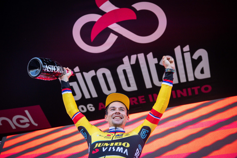 Primož Roglič with his hands up on the podium in front of the Giro d'Italia poster. 