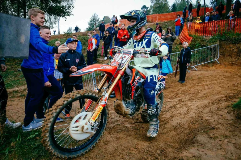 Tim Gajser on his muddy motorcycle in motocross equipment, surrounded by fans on a muddy track.
