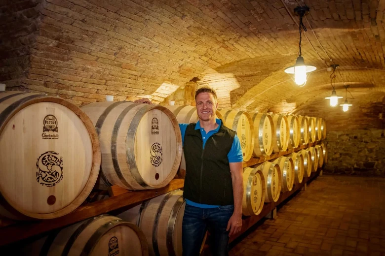 Ralf Schumacher is in a wine cellar, standing next to wooden wine barrels and hugging one barrel.