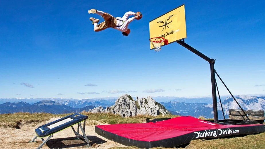 An acrobat from Dunking devils is caught jumping high up in the air, doing a backflip, and about to dunk the basketball in a hoop. There are mountain peaks in the background below.