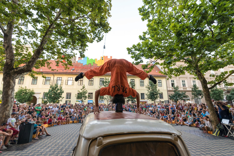 Gledalci na prostem gledajo akrobata, ki na glavi dela stojo na strehi avtomobila.