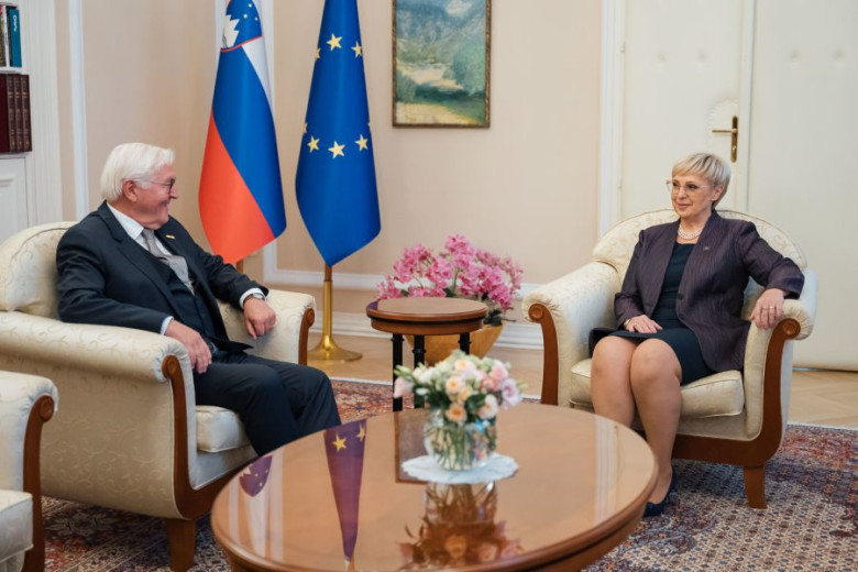 Slovene President Nataša Pirc Musar and German President Frank-Walter Steinmeier meet in a formal sitting room, seated across a flower-topped table with Slovenian and EU flags behind them.