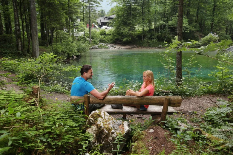A couple sitting on a wooden bench and talking to each other by the Kamniška Bistrica river in a forest. 