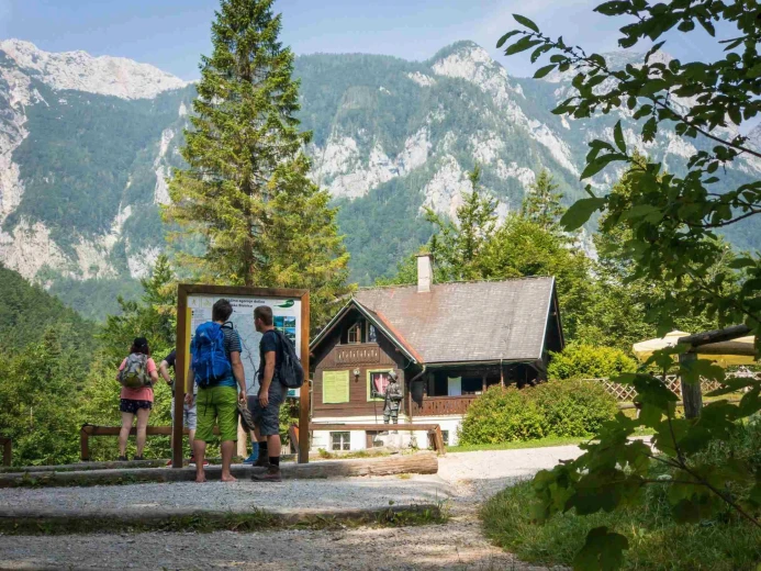 A mountain lodge, surrounded by trees and greenery, and a few hikers in front of the lodge, looking at a map. There are mountains in the background. 