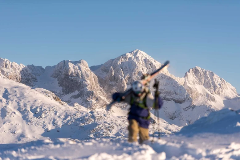 A skier carries skis on his shoulder and walks through the snow. Mountains in the background.