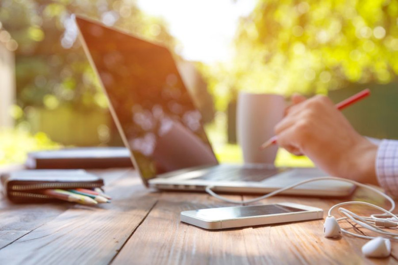 A wooden table sits outdoors with greenery behind it. On the table are a laptop, a mobile phone, and a hand holding a pencil poised above the keyboard.