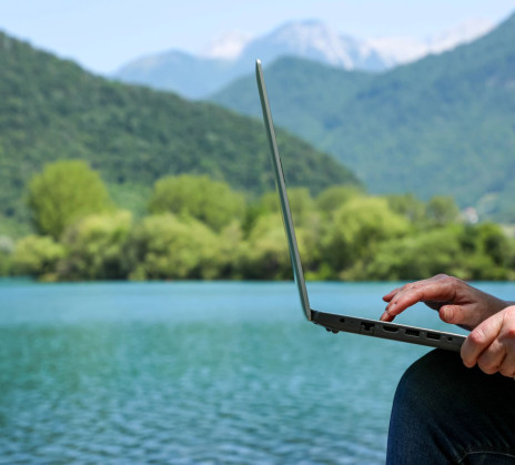 Person using a laptop by a calm lake, with green hills and distant mountains in the background.