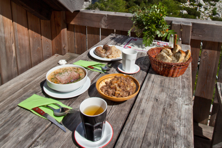 Traditional Slovenian mountain meal served on a wooden outdoor table with a scenic alpine backdrop, including sausage with sauerkraut, jota stew, bread in a basket, and a white mug.