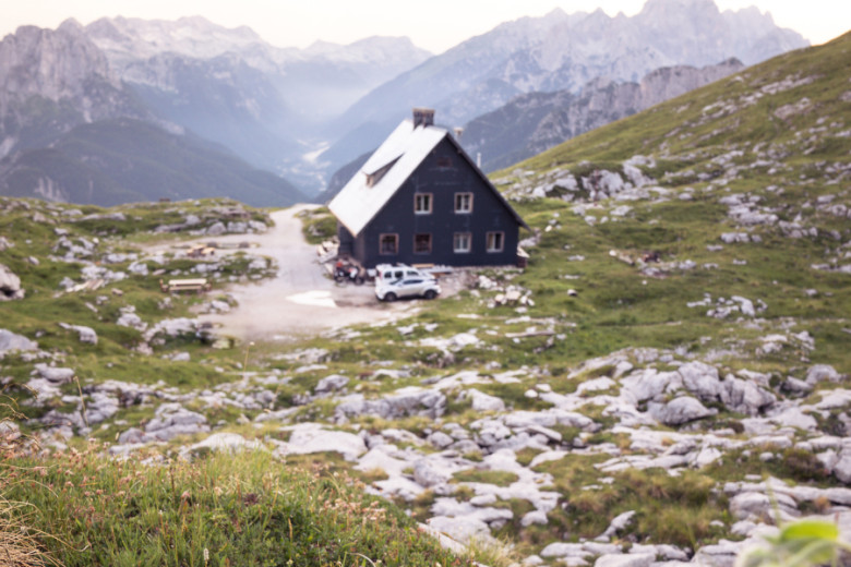 Mountain hut with a steep grey roof and a parked car in front, situated on Mangart Saddle amidst rocky alpine terrain, with dramatic mountain peaks and a misty valley stretching into the distance.