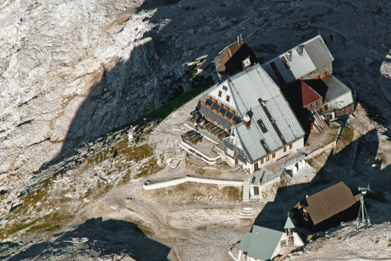 Aerial view of Triglavski dom, a large alpine hut with multiple rooftops, perched on a rocky plateau near the summit of Mount Triglav, surrounded by steep mountain slopes and minimal vegetation.