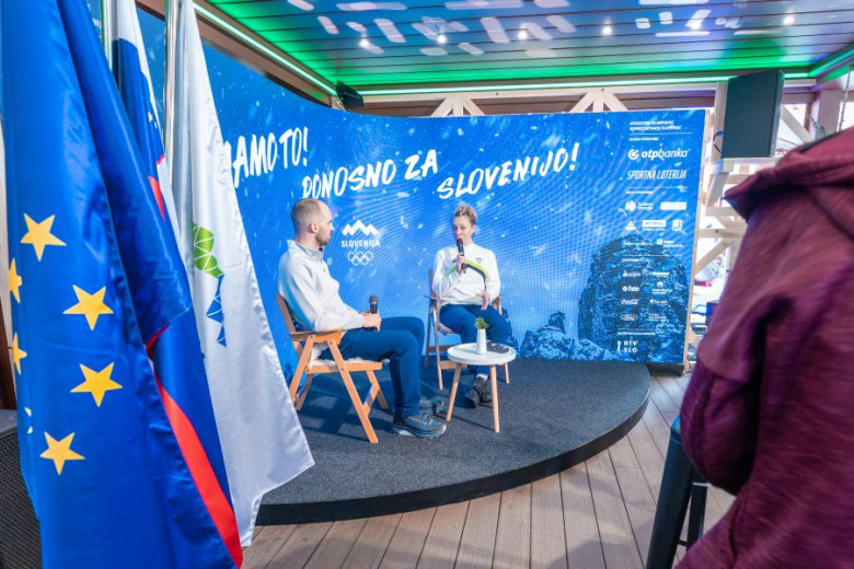 A man and skiier Ilka Štuhec sit on a stage with microphones against a blue backdrop featuring Slovenian Olympic branding.