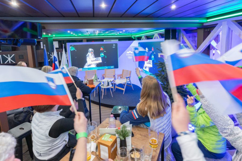 A group of people are gathered around tables wathing a screen, waving Slovenian flags..