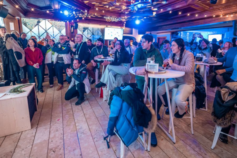 A large group of people in winter attire gathers in a bar and watches a race on screen.