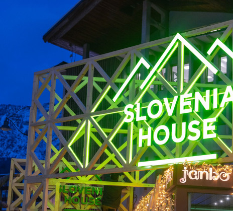 Illuminated sign of "Slovenia House" with green neon lights on a lattice structure against a twilight sky.