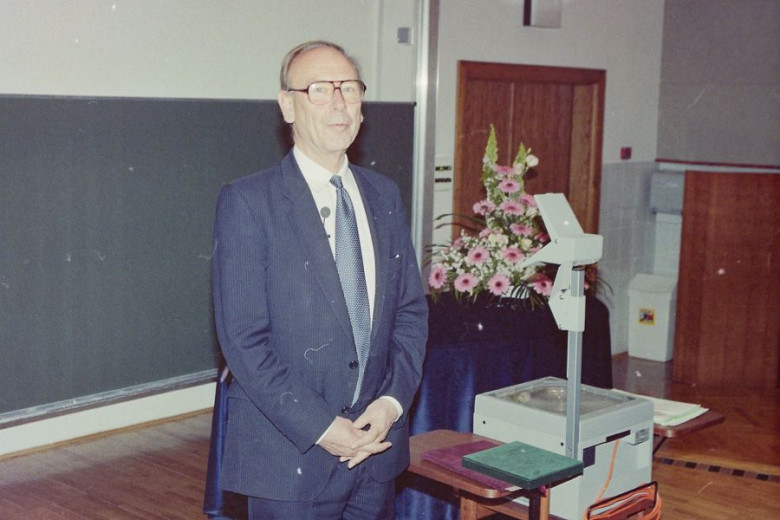 Donald Michie  in a suit stands in a lecture hall beside an overhead projector.
