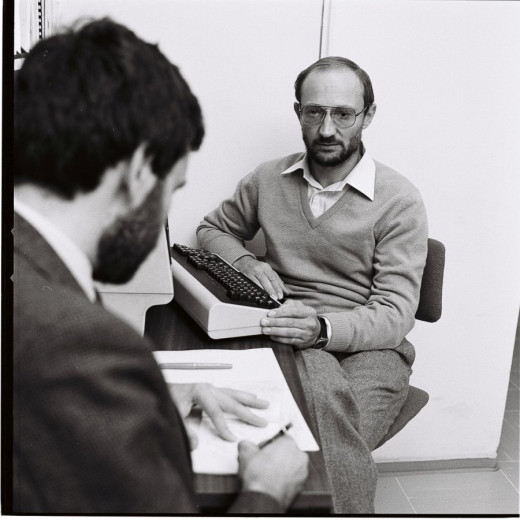 Ivan Bratko with glasses and a sweater, sits beside a computer another man sitting across from him is taking notes.