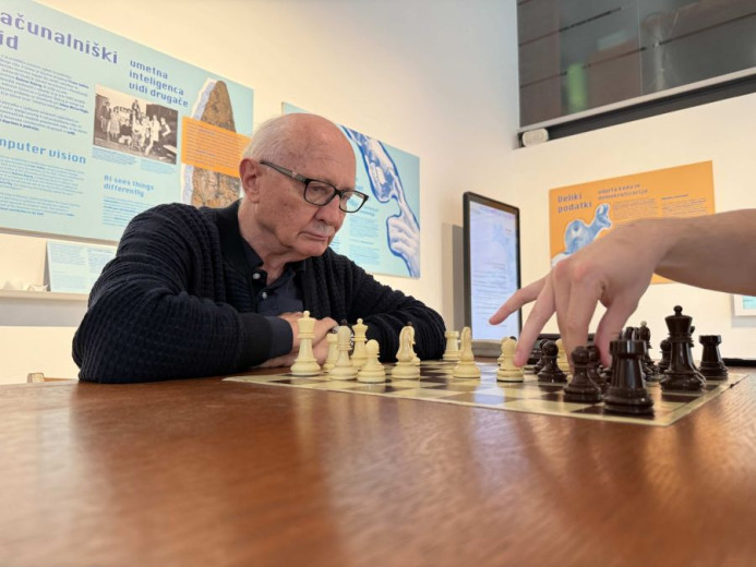 Ivan Bratko wearing glasses is contemplating a chessboard with white pieces. A hand of the other player is moving a black piece.