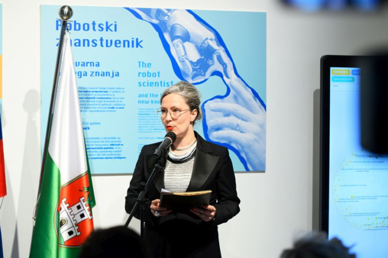 Gaja Zornada in a blazer speaks at a podium with a microphone, holding notes. 
