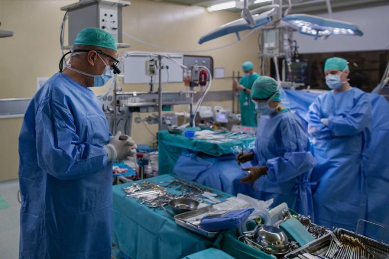 Surgeons in blue scrubs and masks prepare in a brightly lit operating room. Surgical tools are neatly arranged on tables.
