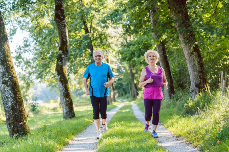 An elderly couple jogs along a tree-lined path on a sunny day.