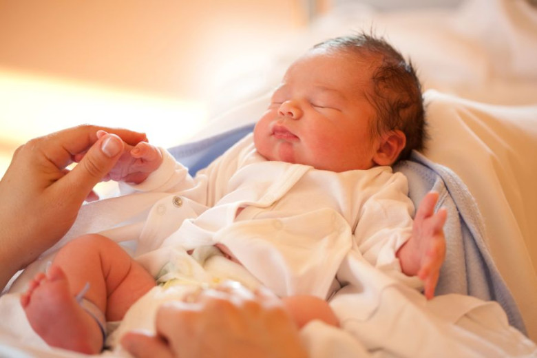 A peaceful newborn baby in a white shirt sleeps in mother’s arms, who gently holds the baby's hand. 