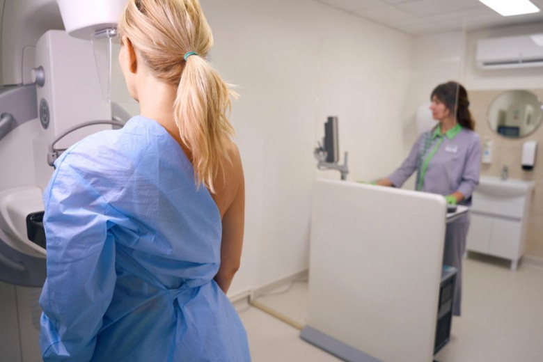 A woman in a blue gown undergoes a mammogram in a medical room. She stands by the machine, with a technician operating equipment nearby. 